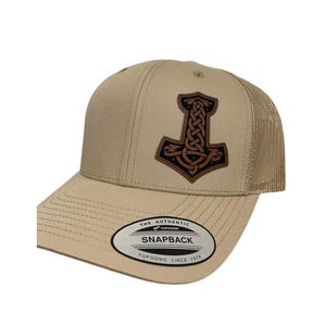 Könnte beinhalten: Eine beige und braune Trucker-Kappe mit einem Leder-Patch, der ein geflochtenes Hammer-Design zeigt. Die Kappe hat ein Netzrücken und einen Druckknopfverschluss. Die Kappe hat einen Aufkleber auf der Vorderseite, auf dem "The Authentic Yupoong Snapback Yupoong Since 1974" steht.