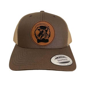 Op de afbeelding: Een bruine en beige trucker-pet met een bruine leren patch met een zwart silhouet van een figuur met een hoed en masker. De patch heeft de tekst "The Authentic Snapback Yupoong Since 1974".
