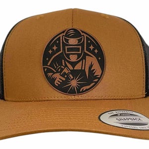 May include: A brown and black snapback cap with a circular emblem of a welder. The cap has a mesh back and a curved peak. The text "THE AUTHENTIC SNAPBACK YUPOONG SINCE 1974" is on the peak.