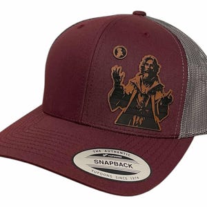 May include: A burgundy and gray trucker hat with a brown leather patch featuring a silhouette of a wizard holding a sphere. The hat has a mesh back and a snap closure. The text "THE AUTHENTIC YUPOONG SNAPBACK YUPOONG SINCE 1974" is printed on the sticker.