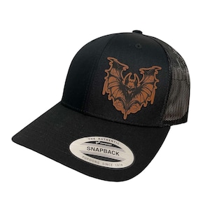 May include: Black and brown baseball cap with a brown leather bat graphic. The cap has a black mesh back and a black snap closure. The cap has a label that reads "The Authentic Yupoong Snapback Yupoong Since 1974".