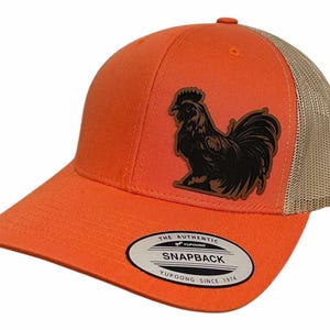 Może przedstawiać: Pomarańczowa i brązowa czapka trucker z czarną naszywką koguta i tekstem "The Authentic Yupoong Snapback Yupoong Since 1974".