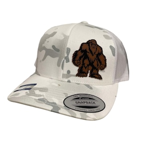 May include: White camouflage trucker hat with a brown leather Bigfoot patch. The hat has a white mesh back and a black snap closure. The hat also has a white label that says "The Authentic Snapback Yupoong Since 1974".
