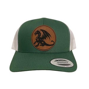 Pode incluir: Boné de caminhoneiro verde com costas de malha branca e um patch de couro marrom com um desenho de dragão preto. O boné tem uma aba curva e um fecho de pressão.