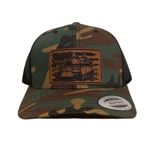 Puede incluir: Una gorra de camionero de camuflaje con un parche de cuero marrón que presenta un tanque con una bandera estadounidense en el fondo. El parche dice "The Authentic Snapback"