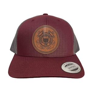May include: A maroon and grey trucker hat with a brown leather patch featuring the United States Coast Guard emblem. The emblem has the words "United States Coast Guard" and "1790" around the outside.