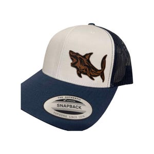 May include: White and navy blue trucker hat with a brown shark graphic and the text "The Authentic Yupoong Snapback Yupoong Since 1974".