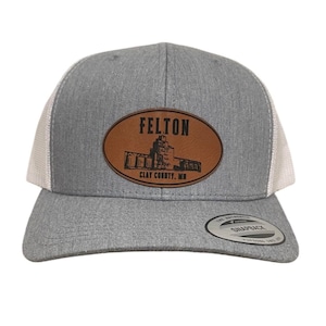 Peut inclure: Casquette de camionneur grise avec un dos en maille blanche et une visière incurvée. L'avant présente un écusson ovale marron avec le mot "FELTON" au-dessus d'une illustration d'un bâtiment et le texte "CLAY COUNTY, MN".