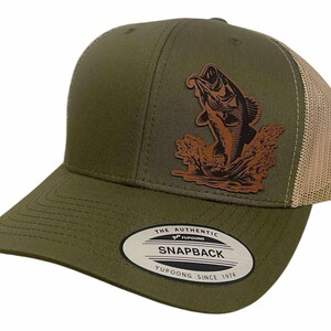 Może przedstawiać: Oliwkowo-zielona i beżowa czapka typu trucker z naszywką ze skóry przedstawiającą wyskakującą rybę. Czapka ma zakrzywiony daszek i siatkową tylną część. Naszywka jest brązowa, a na etykiecie widnieje napis "SNAPBACK".