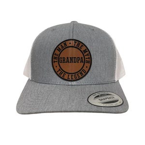 Op de afbeelding: Grijze en witte truckercap met een bruin leren patch waarop staat "The Man, The Myth, The Legend" met "Grandpa" in het midden.