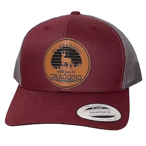 May include: A maroon and gray trucker hat with a brown leather patch that says "No bragging until you're dragging" with a silhouette of a deer. The patch has a sunset graphic with a black background. The hat has a YUPOONG logo on the back.