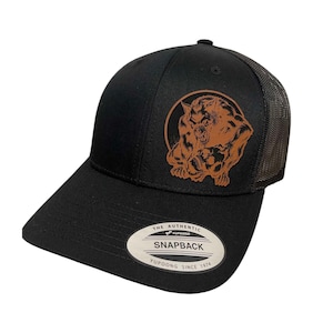 May include: Black and brown trucker hat with a brown leather patch featuring a werewolf illustration. The patch has the text "THE AUTHENTIC SNAPBACK YUPOONG SINCE 1974".