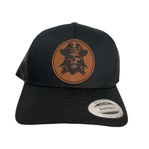 May include: Black trucker hat with a brown leather patch featuring a skull and crossbones pirate design. The patch has the text "The Authentic Snapback Yupoong Since 1974".