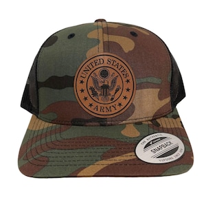 Może przedstawiać: Czapka trucker w kamuflażu z brązową skórzaną naszywką z napisem "United States Army". Czapka ma czarną siateczkową tylną część i białą etykietę z napisem "The Authentic Yupoong Snapback Yupoong Since 1974".