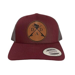 May include: A maroon and gray trucker hat with a brown leather patch featuring a fish and crossed fishing rods with the text "D.D.A."  The hat has a black mesh back and a white label that reads "The Authentic Snapback Trucker Since 1974."
