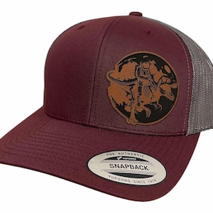 Pode incluir: Boné de caminhoneiro bordô e cinza com um remendo de couro marrom que apresenta um esqueleto de dinossauro em um fundo de lua. O remendo tem o texto "YUPOONG SINCE 1974" e "THE AUTHENTIC SNAPBACK".