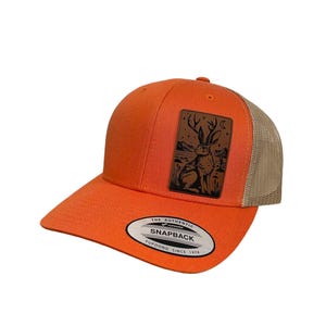 May include: Orange and tan trucker hat with a brown leather patch featuring a black silhouette of a rabbit with antlers. The patch has the text "The Authentic Yupoong Snapback Since 1974".
