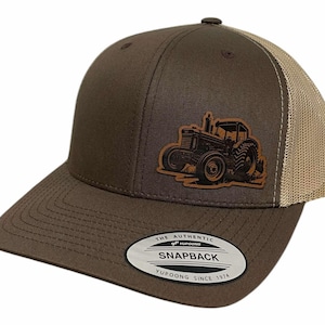 Op de afbeelding: Bruine en beige trucker cap met een bruin leren patch van een tractor op de voorkant. De pet heeft een bruine klep en een beige mesh achterkant. De pet heeft een label op de achterkant met de tekst "The Authentic Yupoong Snapback Since 1974."