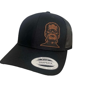 May include: Black snapback hat with a mesh back and a curved brim. The hat features a brown leather patch with a detailed image of Frankenstein's monster. The text "SNAPBACK" and "YUPOONG SINCE 1974" are visible on the front.