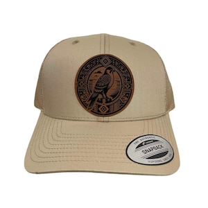 Peut inclure: Un chapeau de camionneur beige avec un patch en cuir marron représentant un faucon stylisé. Le patch présente un motif géométrique autour du faucon. Le chapeau a un dos en maille et le texte "The Authentic Snapback Yupoong Since 1974" au dos.