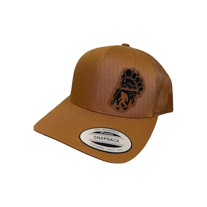 Op de afbeelding: Een bruine en zwarte trucker-pet met een leren patch met een voetafdruk en een Bigfoot-silhouet. De patch heeft de tekst "The Authentic" en "Snapback" erop. De pet heeft een mesh-rug.