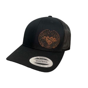 May include: Black and brown trucker hat with a brown graphic of a bat with a moon and trees. The hat has a black mesh back and a black snap closure. The hat has a label that says "The Authentic Yupoong Snapback Since 1974."