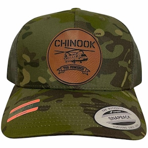 May include: Camouflage pattern baseball cap with a brown leather patch featuring the word "CHINOOK" and a helicopter graphic. The cap has a mesh back and a curved brim. The hat is a snapback style.