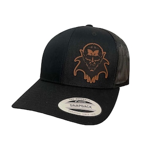 Puede incluir: Gorra de camionero negra y marrón con un parche de cuero que presenta una cara de vampiro. La gorra tiene una parte trasera de malla negra y un cierre de broche negro. La gorra también tiene una etiqueta que dice "The Authentic Yupoong Snapback Yupoong Since 1974."