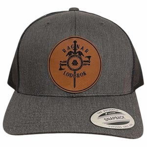May include: A gray trucker hat with a black mesh back and a brown leather patch. The patch features the words "RAGNAR LODBROK" around a Viking-themed design. The hat has a curved brim and a snapback closure.