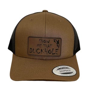 Puede incluir: Una gorra de camionero marrón y negra con un parche de cuero marrón que dice "Show Me That Duckhole". La gorra tiene una parte trasera de malla negra y un cierre de presión YUPOONG marrón.