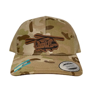 Puede incluir: Una gorra de camionero de camuflaje marrón con un parche de cuero marrón de un helicóptero Chinook en la parte delantera. La gorra tiene una parte trasera de malla negra y un cierre de presión negro. La gorra también tiene una pequeña etiqueta azul y blanca que dice "MULTICAM" en el lateral. La gorra tiene una etiqueta blanca en la parte posterior que dice "THE AUTHENTIC SNAPBACK YUPOONG SINCE 1974."