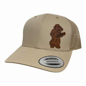 Könnte beinhalten: Beige Baseballcap mit Mesh-Rückseite und gebogenem Schirm. Das Cap hat einen Lederaufnäher mit einem Cartoon-Alligator, der einen Hut trägt und ein Gewehr hält. Auf der Vorderseite steht "Snapback" und "Yupoong Since 1974".