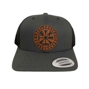 May include: A dark gray trucker hat with a black mesh back. The hat features a brown leather patch with a black compass design and runes around the edge. The text "THE AUTHENTIC SNAPBACK YUPOONG SINCE 1974" is on a white tag attached to the hat.