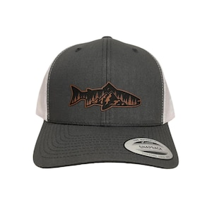 May include: A dark grey and white trucker hat with a brown leather patch of a fish silhouette with mountains and trees inside the fish. The text "The Authentic Snapback" is on the back of the hat.