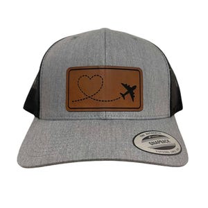 May include: Gray trucker hat with a black mesh back and a curved brim. A brown leather patch features a heart-shaped dotted line and a black airplane graphic. The hat has a snapback closure.