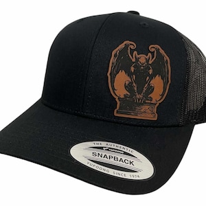 Könnte beinhalten: Schwarze und braune Trucker-Kappe mit einem Leder-Patch, der eine Gargoyle mit Flügeln zeigt. Die Kappe hat ein schwarzes Netzrücken und eine schwarze Vorderseite mit einem weißen Etikett, auf dem "The Authentic Yupoong Snapback Yupoong Since 1974" steht.