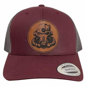 Puede incluir: Gorra de camionero burdeos y gris con un parche de cuero que presenta una ilustración detallada de un casco futurista. La gorra tiene una visera curva y un cierre a presión. El parche es circular y parece ser de cuero marrón.
