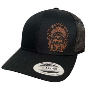 May include: Black trucker hat with a brown leather patch featuring a Native American headdress with a skull. The hat has a black mesh back and a black snap closure. The text "The Authentic Yupoong Snapback Yupoong Since 1974" is printed on the label.