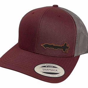 May include: A maroon and gray trucker hat with a brown leather patch of a drone silhouette. The hat has a "The Authentic Yupoong Snapback" label on the back.