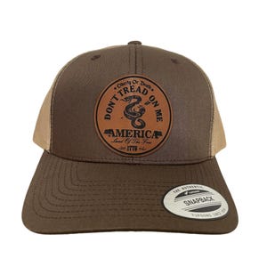 May include: A brown and tan trucker hat with a brown leather patch that says "Don't Tread On Me" with a snake and "America Land of the Free 1776" in a circle.