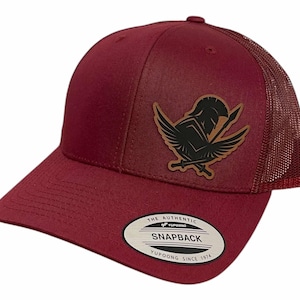 Könnte beinhalten: Eine bordeauxrote Trucker-Kappe mit einem braunen Leder-Patch, der einen geflügelten spartanischen Krieger mit Speer und Schwert zeigt. Die Kappe hat ein Netzrücken und ein "The Authentic Yupoong Snapback Yupoong Since 1974"-Etikett.