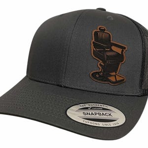 May include: A gray and black trucker hat with a brown leather patch of a barber chair. The hat has a black mesh back and a gray front with the text "The Authentic Yupoong Snapback Yupoong Since 1974" on the back.