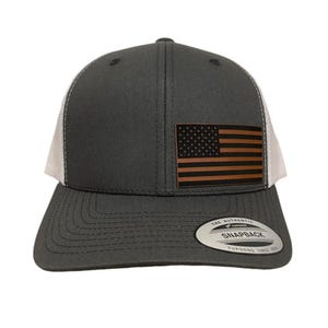 May include: A dark grey and white trucker cap with a brown leather American flag emblem. The cap has a curved peak and a snapback fastening. The tag reads "THE AUTHENTIC SNAPBACK YUPOONG SINCE 1974."