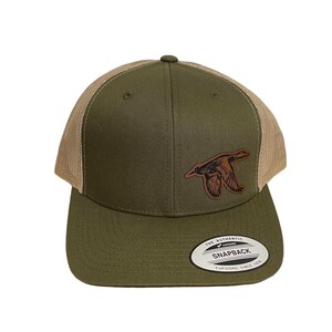 Puede incluir: Una gorra de camionero verde y marrón con un diseño de pato de cuero marrón. La gorra tiene una parte trasera de malla y una visera curvada. La gorra tiene una etiqueta que dice "The Authentic Yupoong Snapback Yupoong Since 1974".