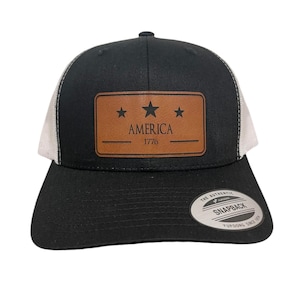 May include: A black and white trucker hat with a brown leather patch that says "AMERICA 1776" with three stars. The hat has a white mesh back and a black front.