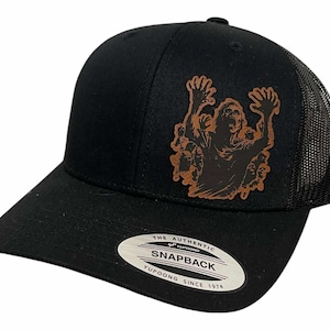 Peut inclure: Casquette de camionneur noire avec un patch en cuir marron représentant une figure stylisée aux bras levés. La casquette a une visière incurvée et un dos en filet. Le texte "THE AUTHENTIC SNAPBACK YUPOONG SINCE 1974" est sur le devant.