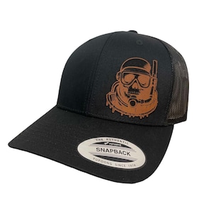 May include: Black and brown trucker hat with a leather patch of a skull wearing a scuba mask and diving gear. The hat has a black mesh back and a white label that reads "The Authentic Yupoong Snapback Yupoong Since 1974".