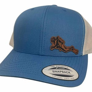 May include: Blue and tan Snapback hat with a diver patch. The hat features a blue front panel and bill, with a tan mesh back. A brown leather patch of a diver is on the front. The hat has a Snapback closure and a Yupoong tag.