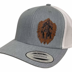 May include: A gray and white trucker hat with a brown leather patch featuring a skull and crossbones design. The patch has the text "The Authentic Yupoong Snapback Yupoong Since 1974".