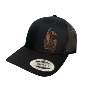 Peut inclure: Casquette de baseball noire Snapback avec un motif de pirate marron. La casquette a une visière incurvée, un dos en filet et le texte "THE AUTHENTIC SNAPBACK YUPOONG SINCE 1974" sur un patch blanc.